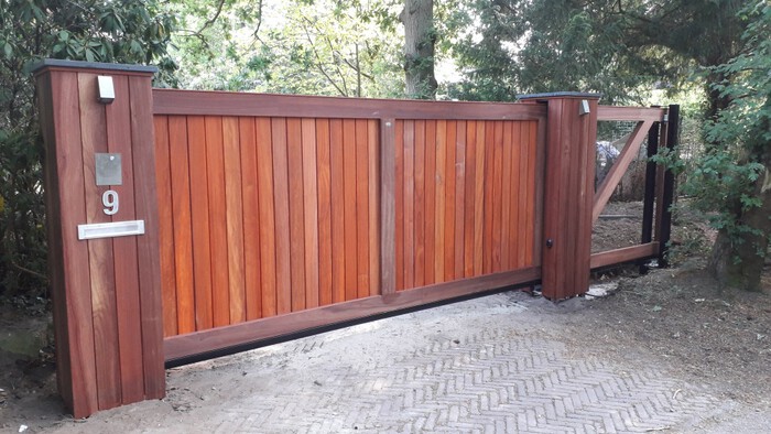 Houten schuifpoort met verticale hardhouten delen tussen twee gemetselde poortpalen aan een bosrijke oprit